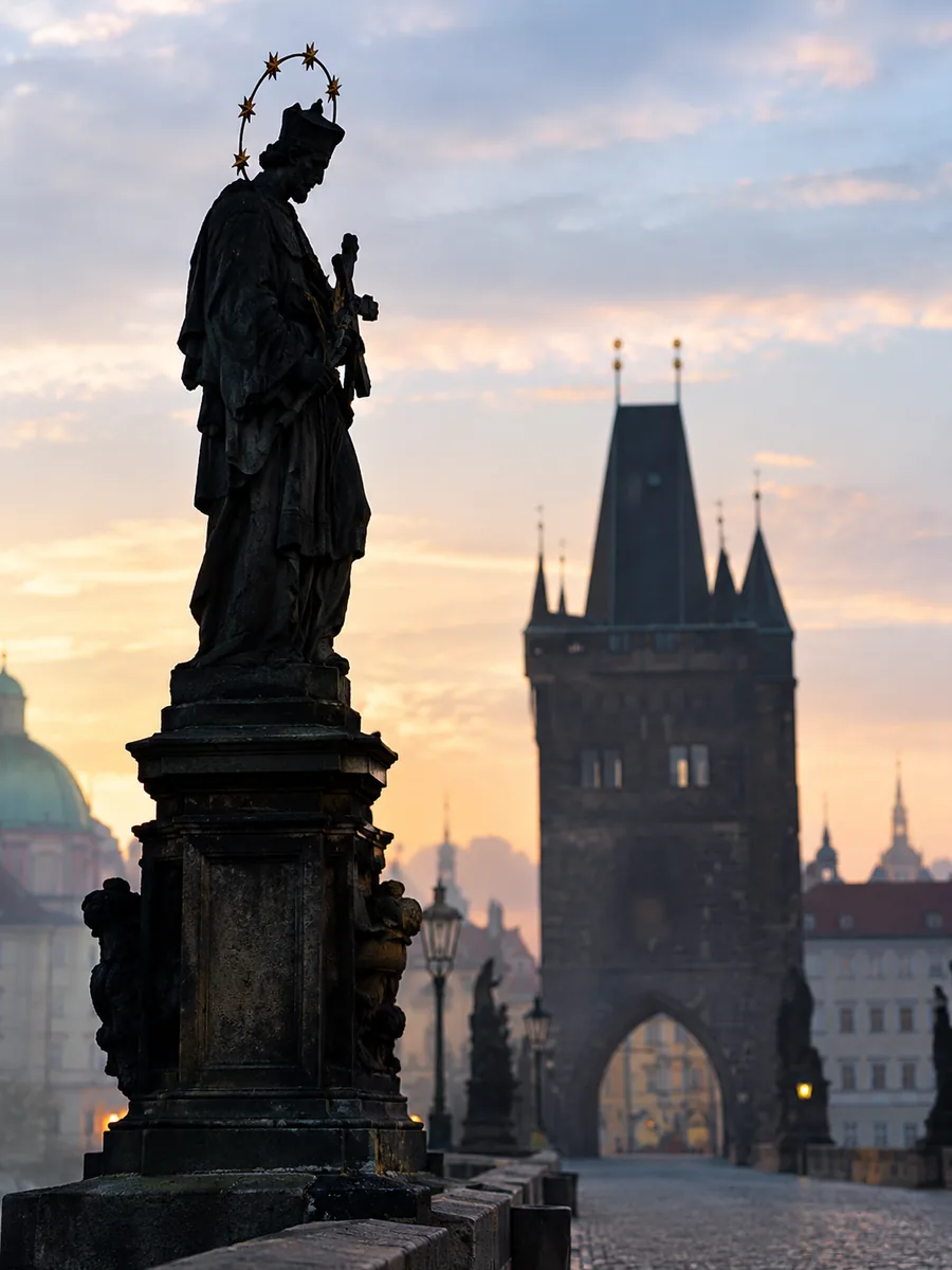 Illustration einer barocken Heiligenstatue auf der Karlsbrücke, silhouettiert gegen den Morgenhimmel