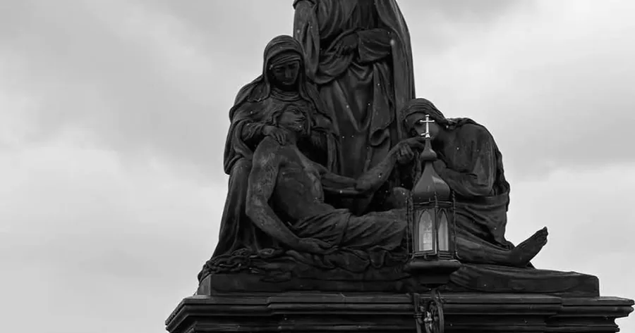 A dark statue group on Charles Bridge in black and white, weathered sandstone against a pale sky